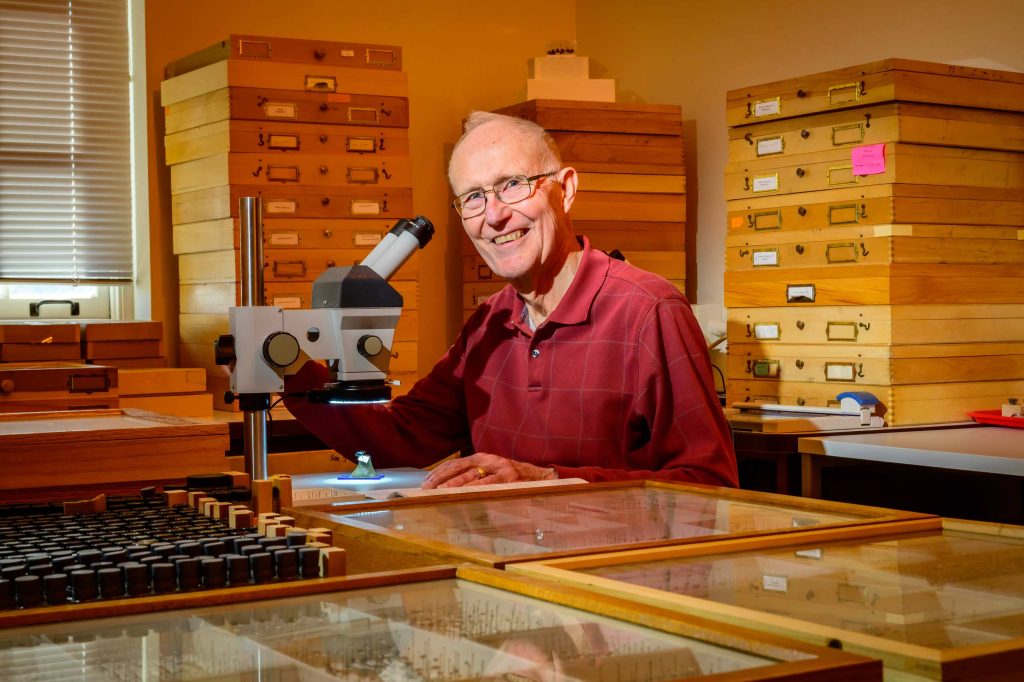 Smiling man with microscope sits among piles of wooden insect drawers.